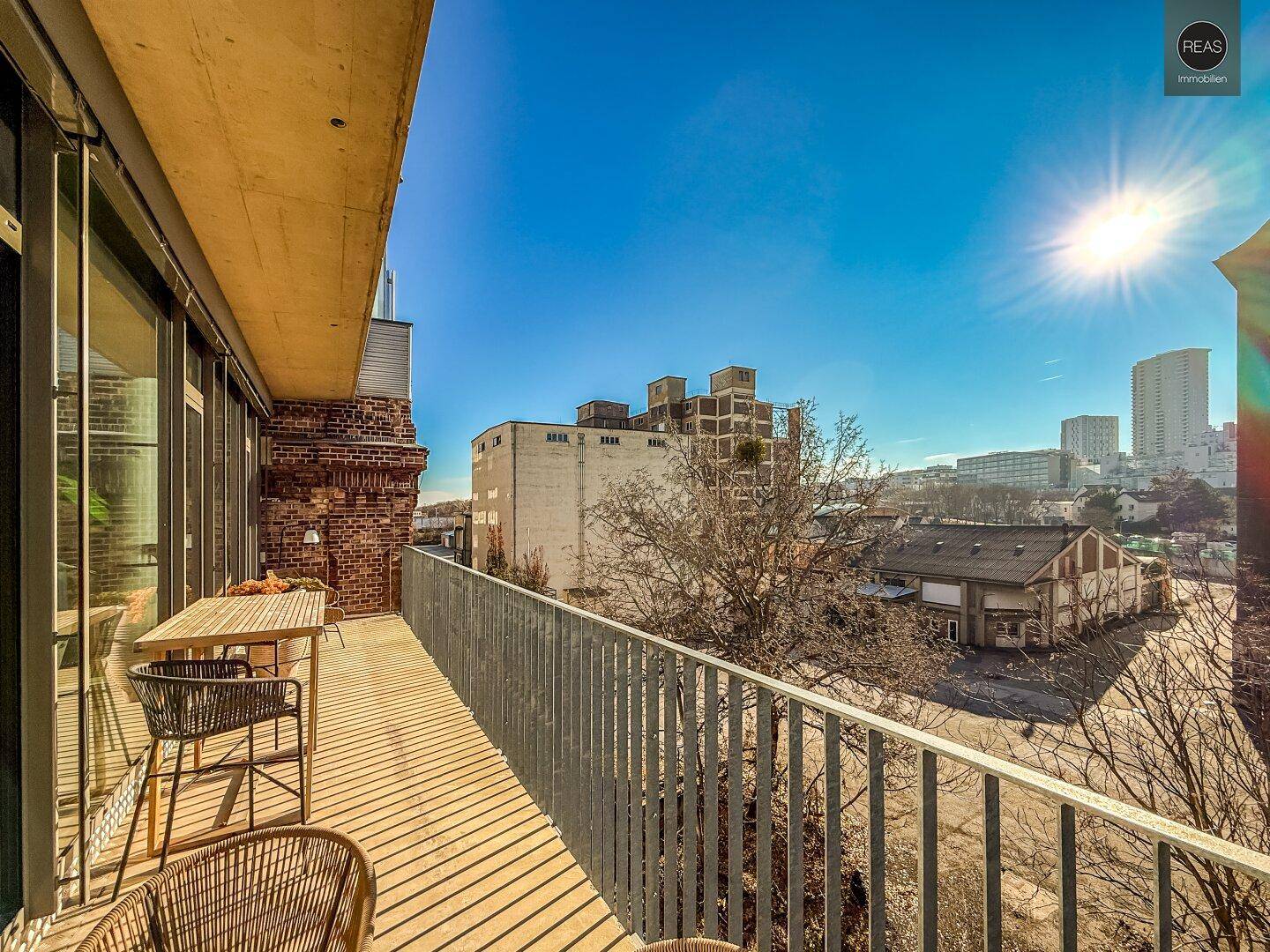 Langer Balkon mit Sitzgelegenheiten und Blick auf die Stadt und umliegende Gebäude unter strahlendem Sonnenschein.