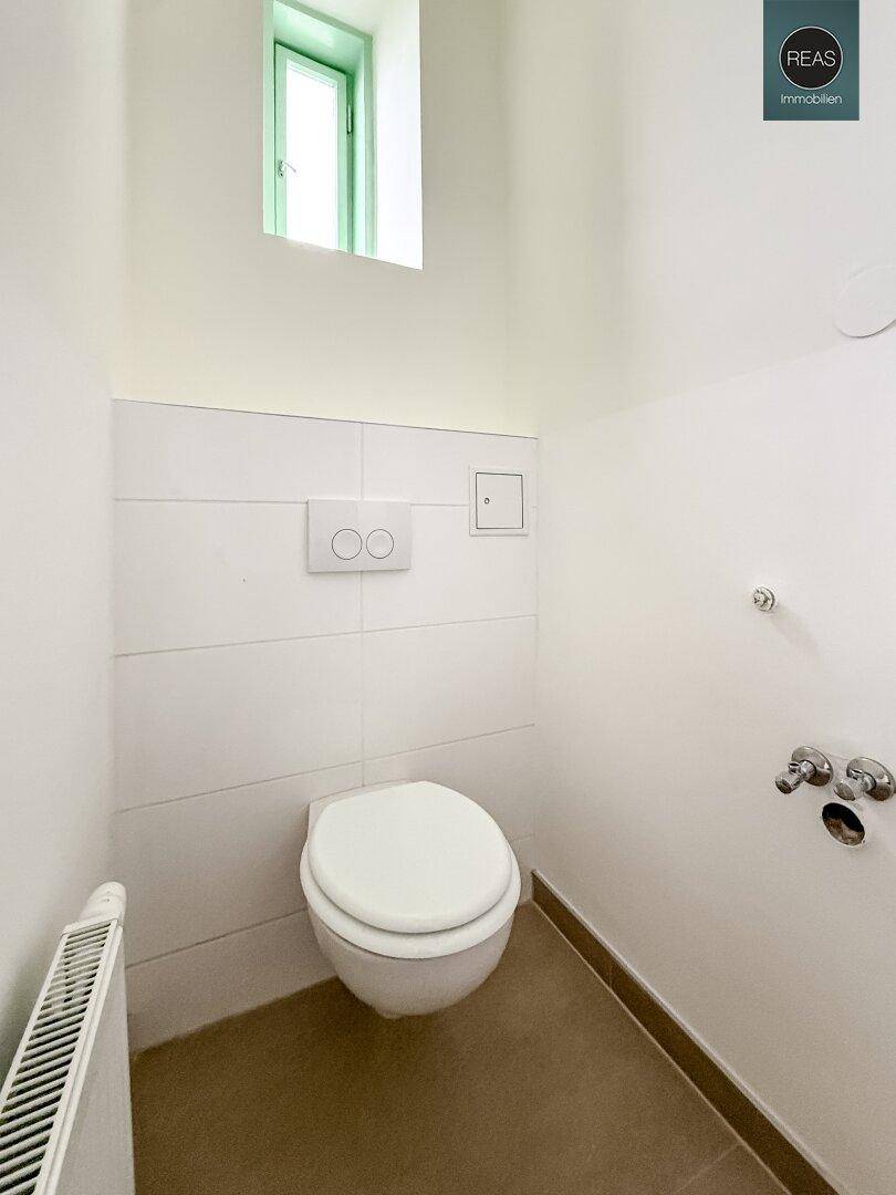 Separate Toilette mit Wand-WC, Heizkörper und einem kleinen Fenster, hell gefliest.