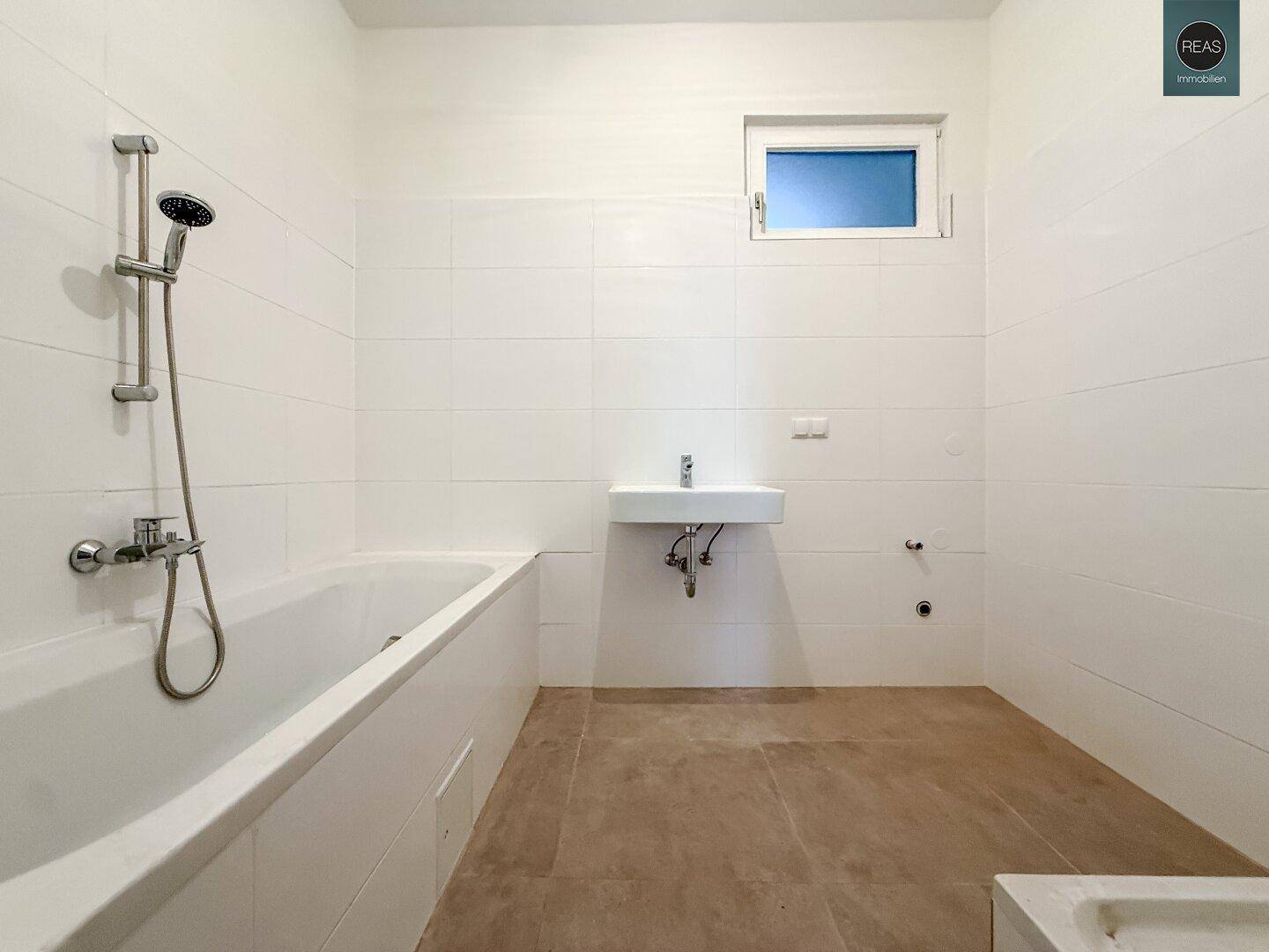 Helles Badezimmer mit Badewanne, Duscharmatur, Waschbecken und einem kleinen Fenster.