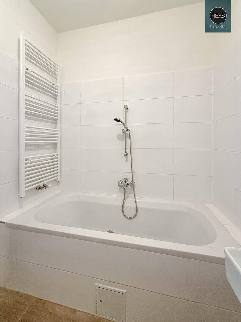 Badezimmer mit Badewanne, Duscharmatur und Handtuchheizkörper, gefliest in Weiß und Beige.