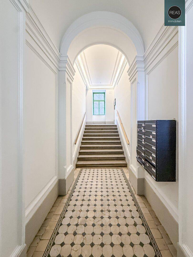 Langer Flur mit hohen Decken, Bogen und gemusterten Fliesen, führt zu einer Treppe.