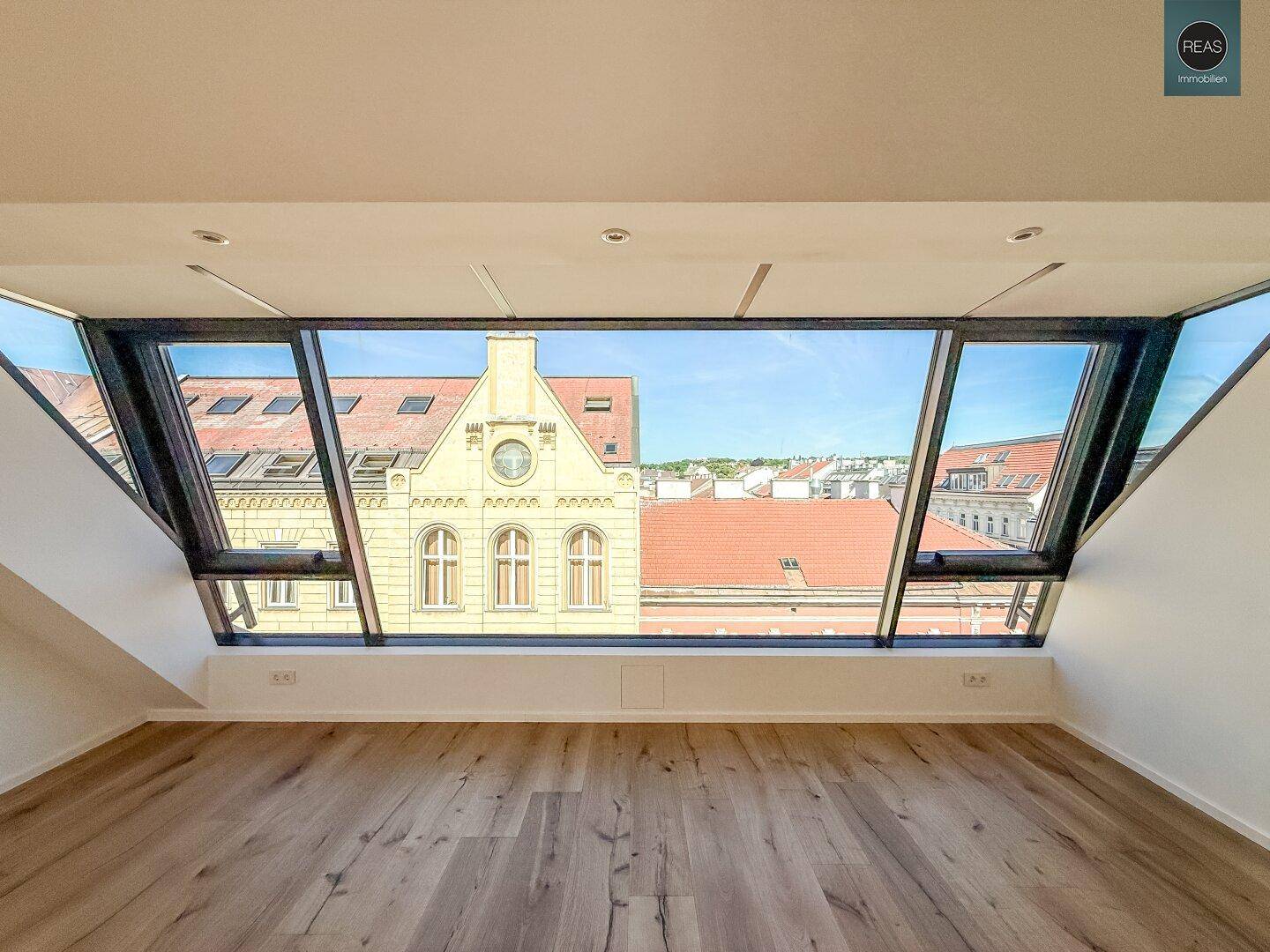 Heller Raum mit Holzboden und großen Dachfenstern, die einen Blick auf historische Gebäude ermöglichen.