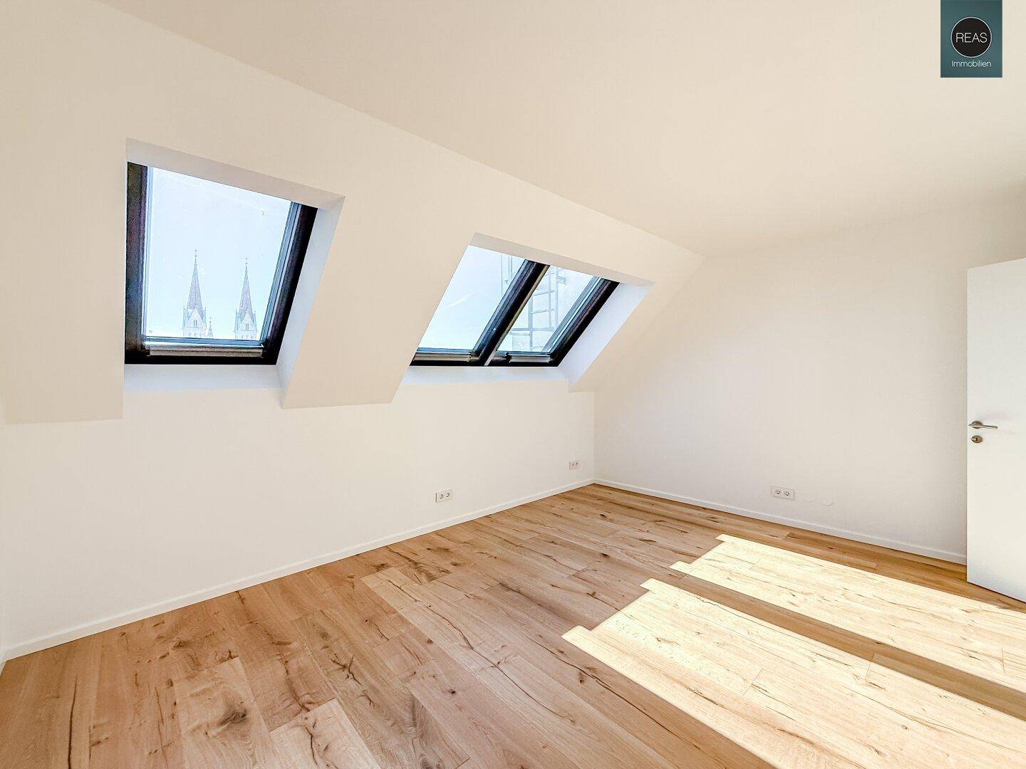Helles Zimmer mit Holzboden und zwei Dachfenstern, die einen Blick auf Kirchtürme bieten.