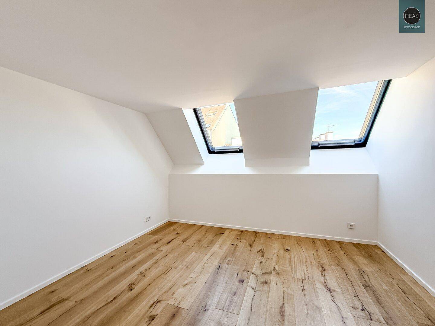 Helles Zimmer mit Holzboden und zwei Dachfenstern, die viel Tageslicht hereinlassen.