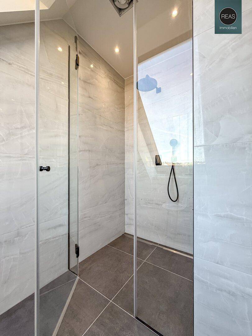 Moderne begehbare Dusche mit Glasabtrennung und Regenduschkopf in einem gefliesten Bad.