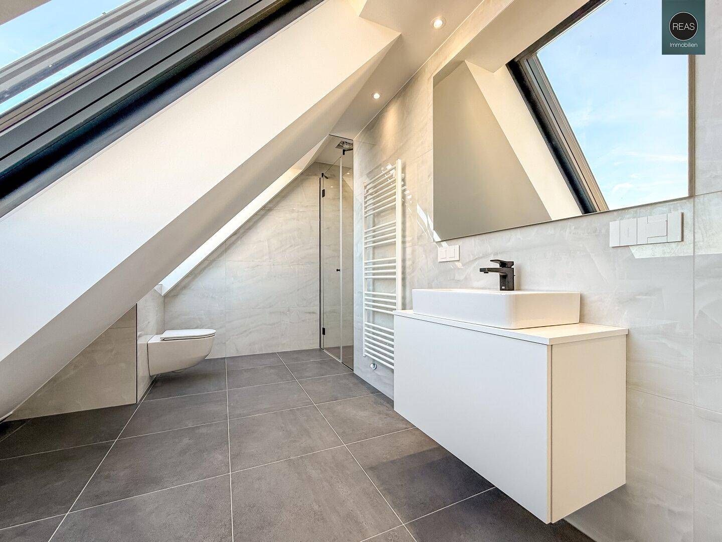 Modernes Badezimmer mit grauen Fliesen, wandhängendem WC und begehbarer Dusche.