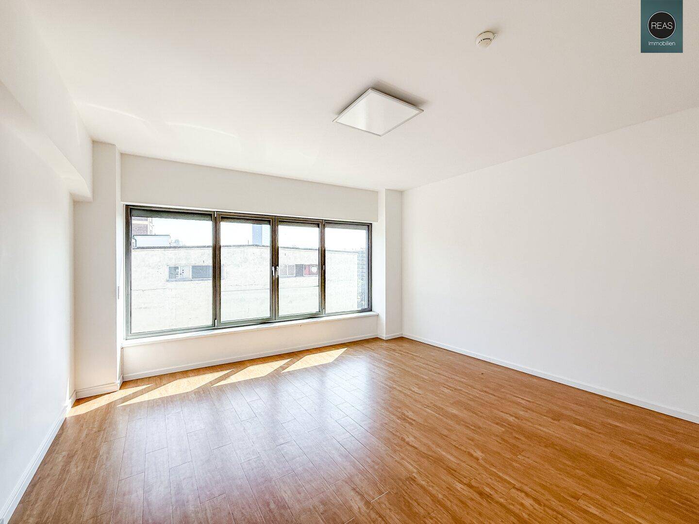 Heller, leerer Büroraum mit großen Fenstern, die viel Tageslicht hereinlassen, und Holzboden.