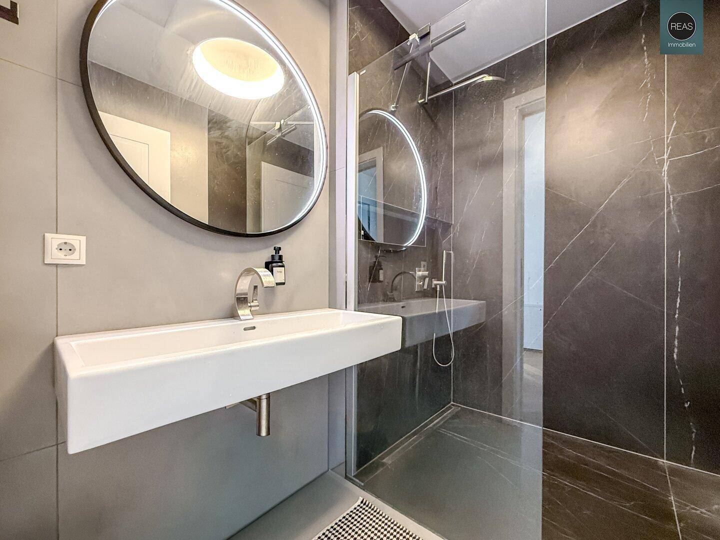 Modernes Badezimmer mit großer Dusche, eleganten Fliesen und ovalem Spiegel.