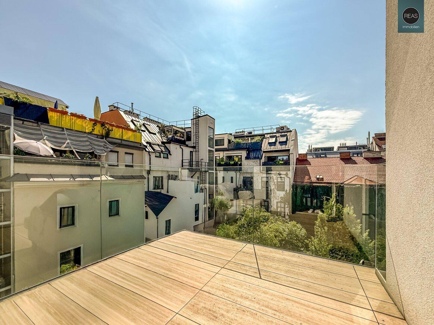 Sonnige Dachterrasse mit Holzboden und Glasgeländer, umgeben von städtischen Gebäuden.
