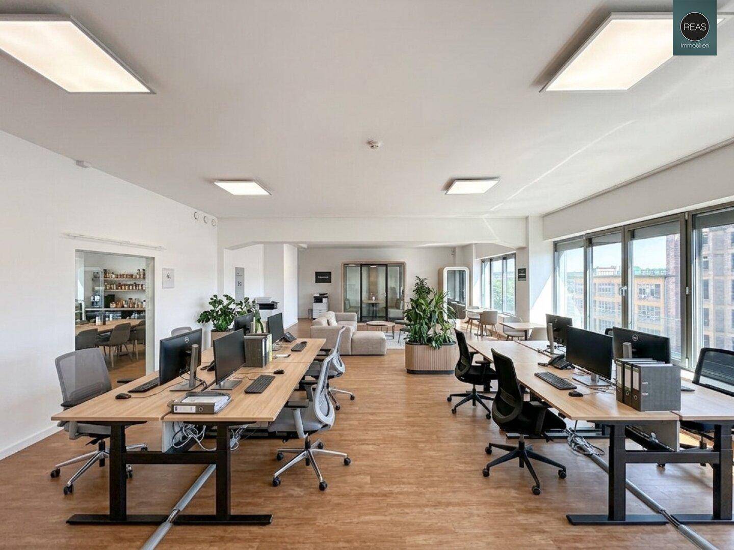 Moderner Büroraum mit mehreren Arbeitsplätzen, Computern, Pflanzen und großen Fenstern mit Stadtblick.