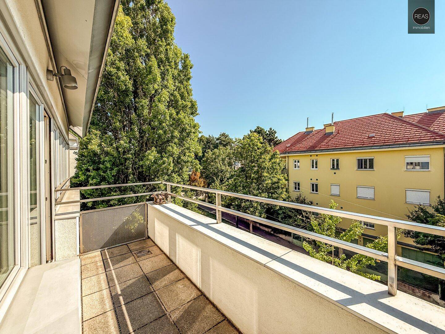 Langer Balkon mit Metallgeländer und Blick auf die grünen Bäume und umliegenden Gebäude.