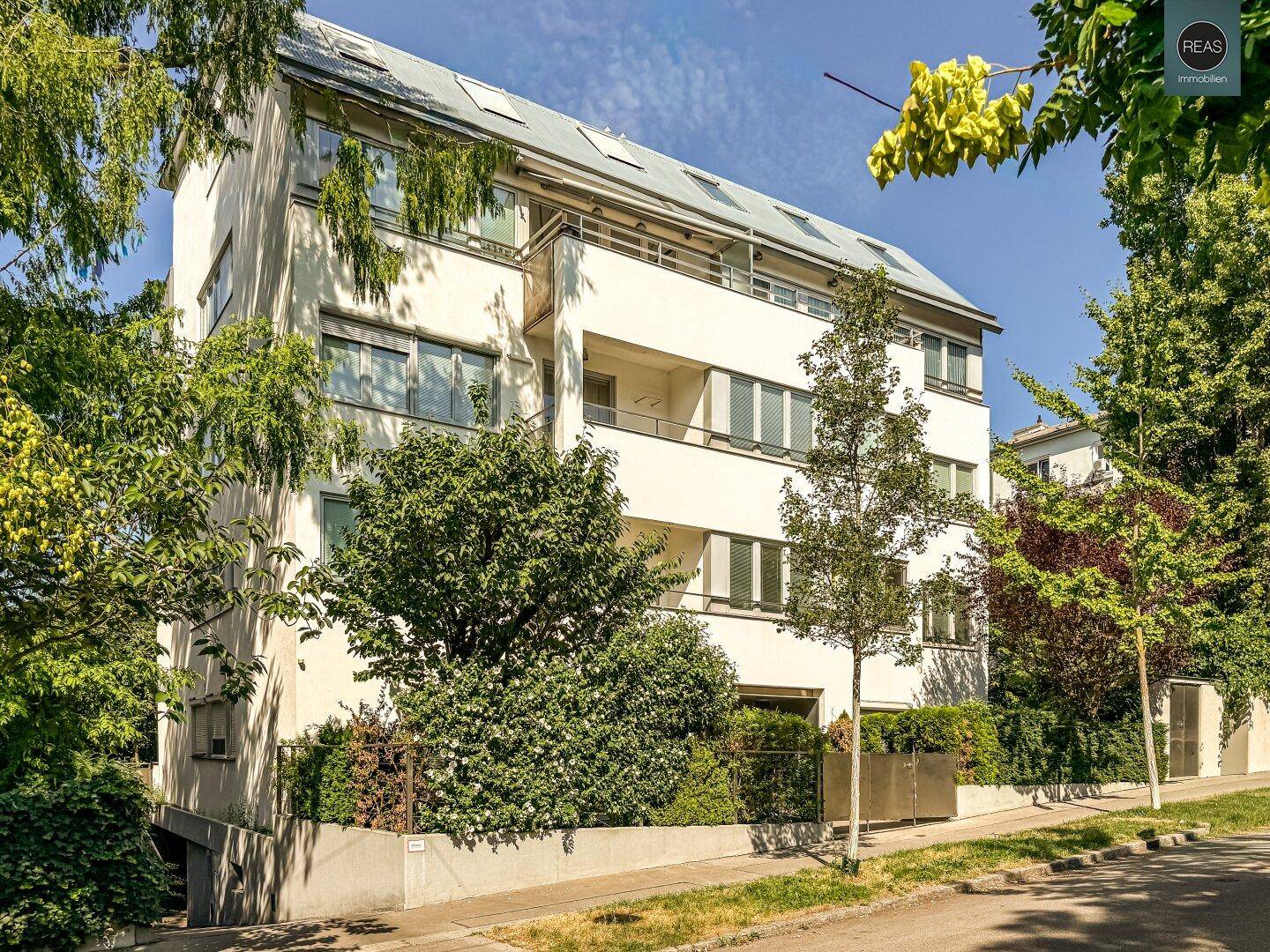 Mehrfamilienhaus mit weißer Fassade, Balkonen und vielen Bäumen in der Umgebung.