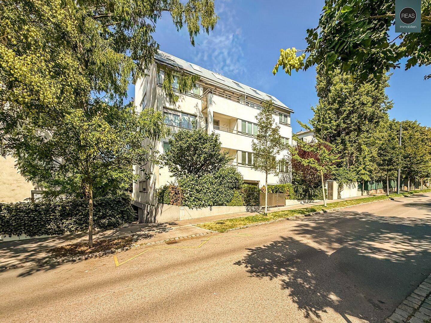 Wohngebäude an einer ruhigen Straße, umgeben von viel Grün und Bäumen unter blauem Himmel.
