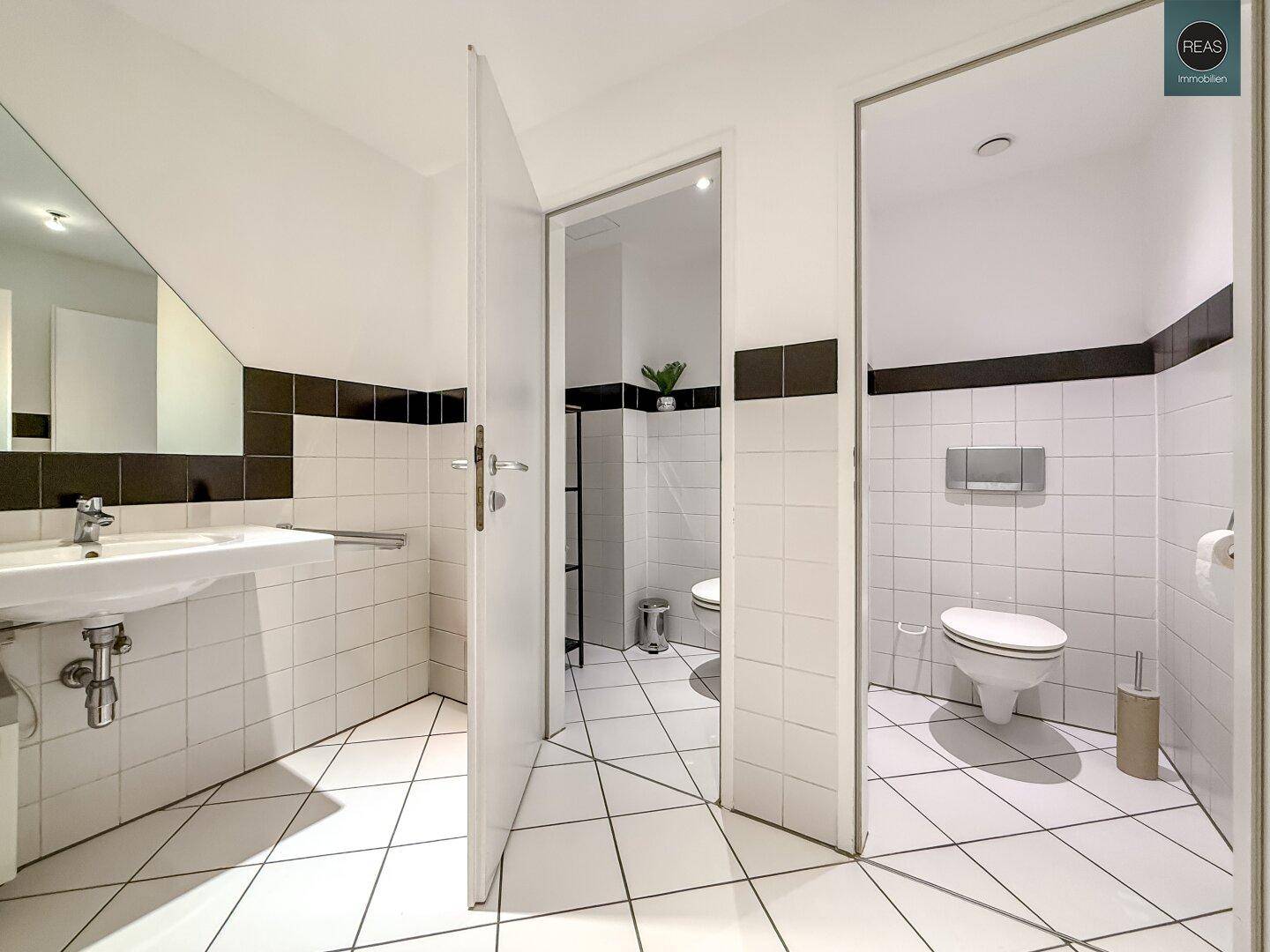 Helles Badezimmer mit weißen Fliesen, schwarzen Akzenten und zwei separaten Toiletten.