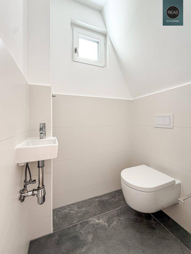 Helles Gäste-WC mit wandhängendem WC, kleinem Waschbecken und Dachfenster für Belüftung.