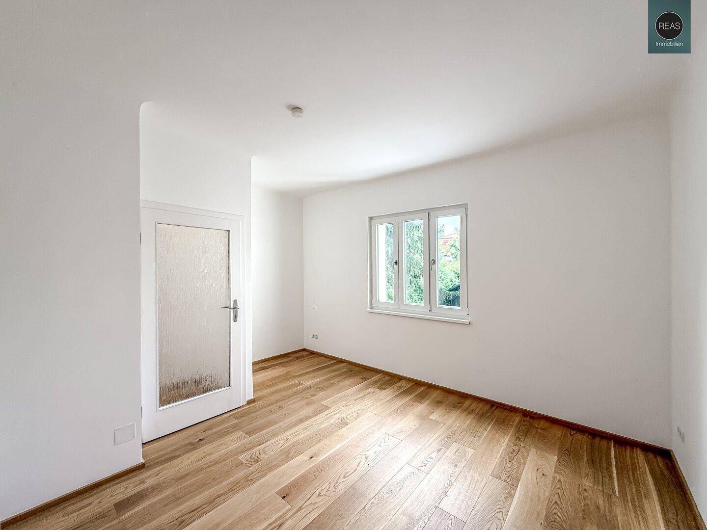Helles, leeres Zimmer mit Eichenparkettboden und einem großen Fenster mit Blick ins Grüne.