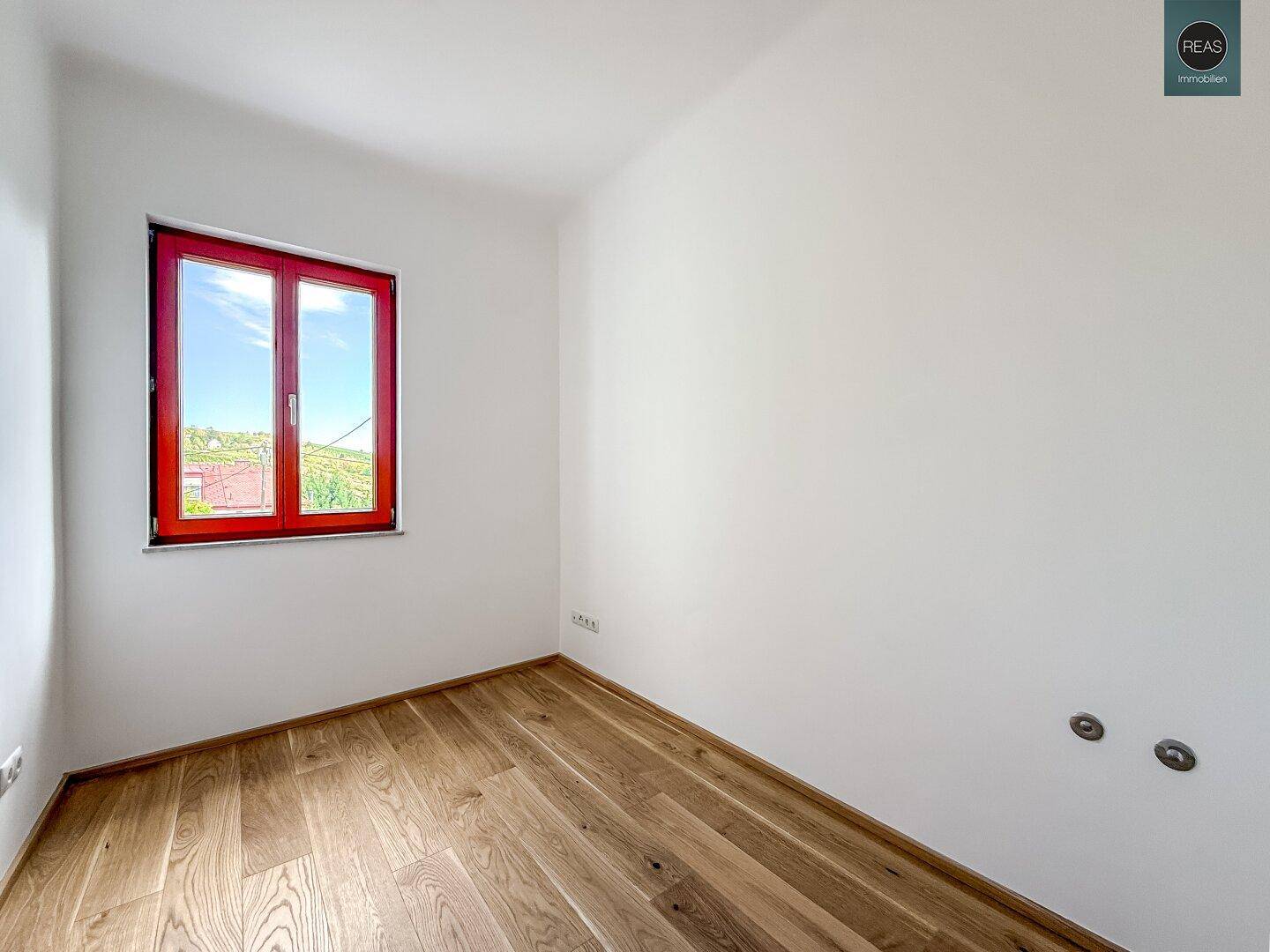 Helles Zimmer mit Holzboden und rotem Fensterrahmen, ideal als Schlaf- oder Arbeitszimmer.