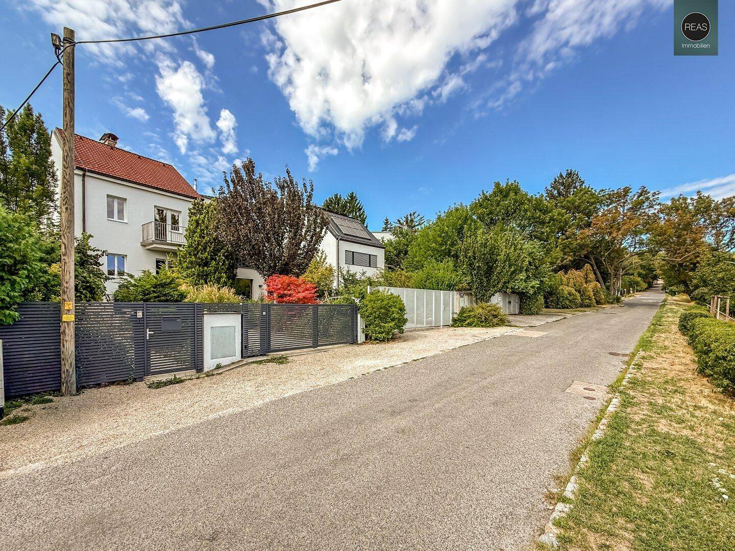 Ansicht des Hauses von der Straße mit Zaun, Einfahrt und umliegender Vegetation.
