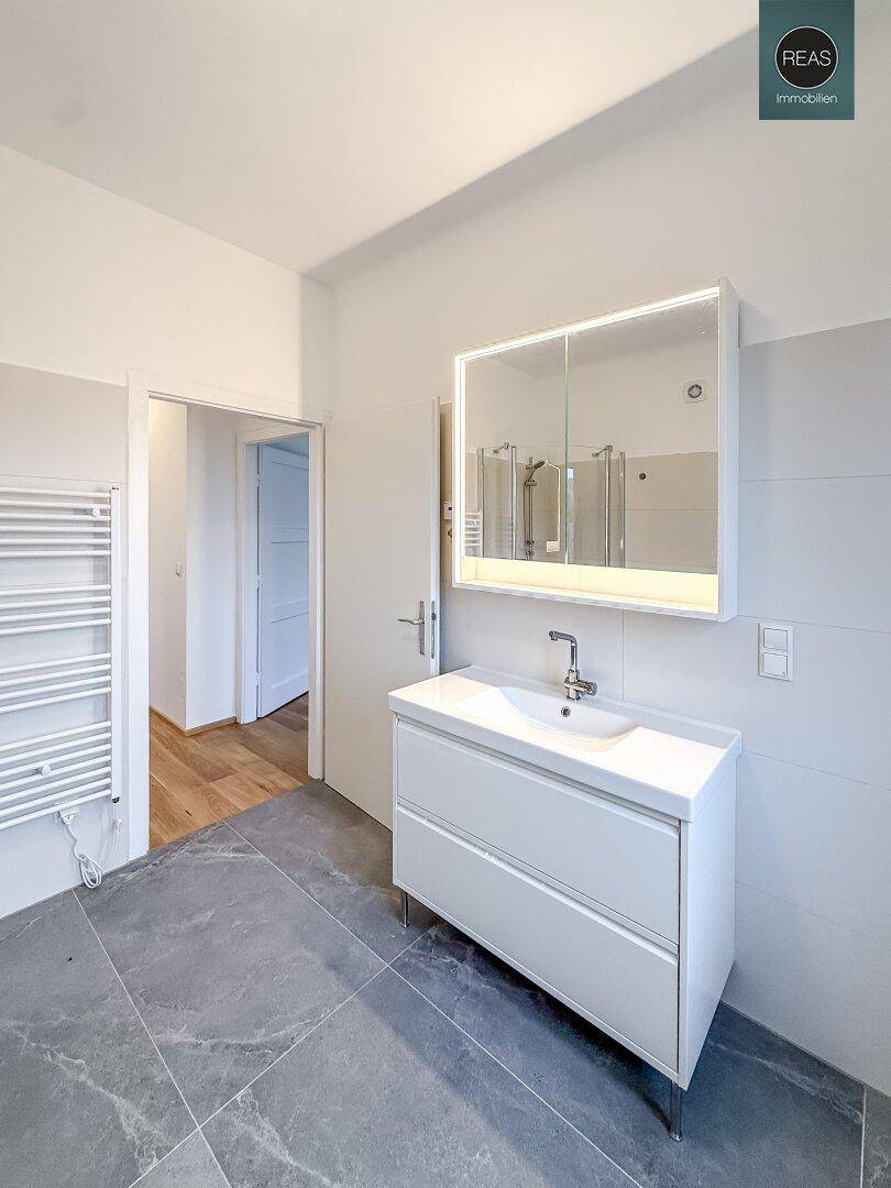 Modernes Badezimmer mit Waschtisch, beleuchtetem Spiegelschrank, großer Dusche und grauen Fliesenboden.