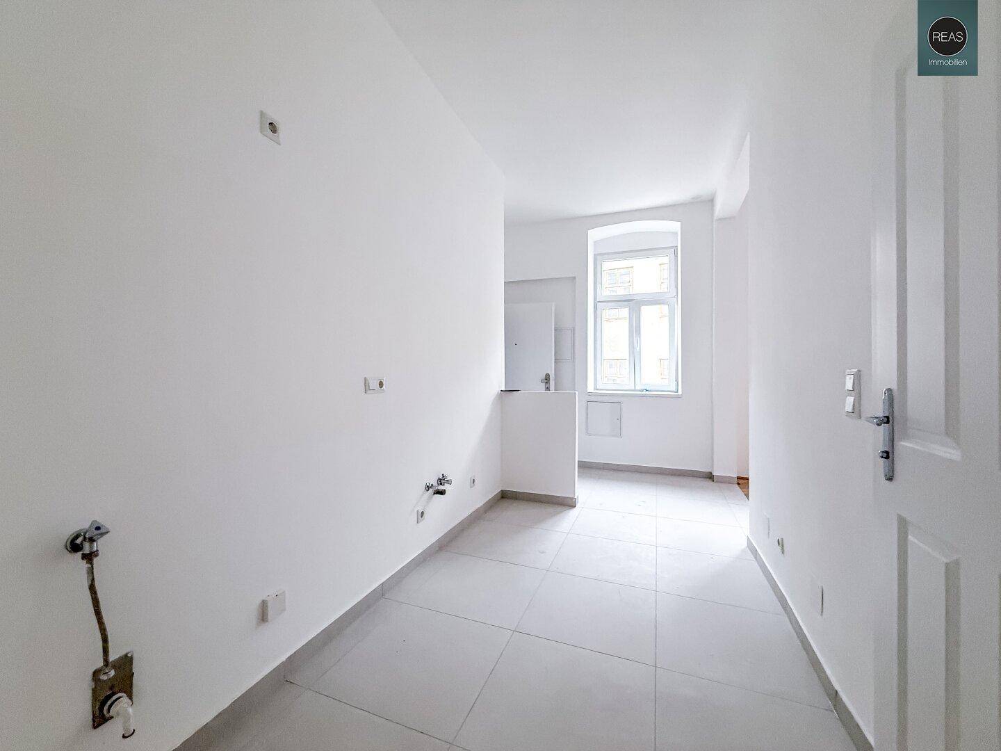 Langer Raum mit Fliesenboden und Fenster, vorbereitet für die Kücheninstallation mit sichtbaren Anschlüssen.