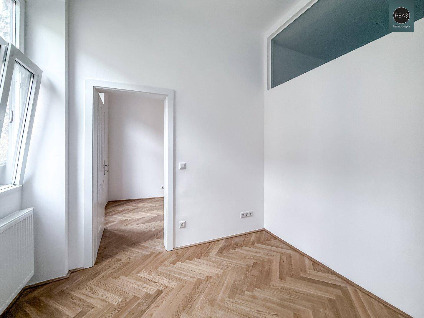 Heller Raum mit Fischgrätparkett und einem hohen Fenster, das viel Licht in den Bereich bringt.