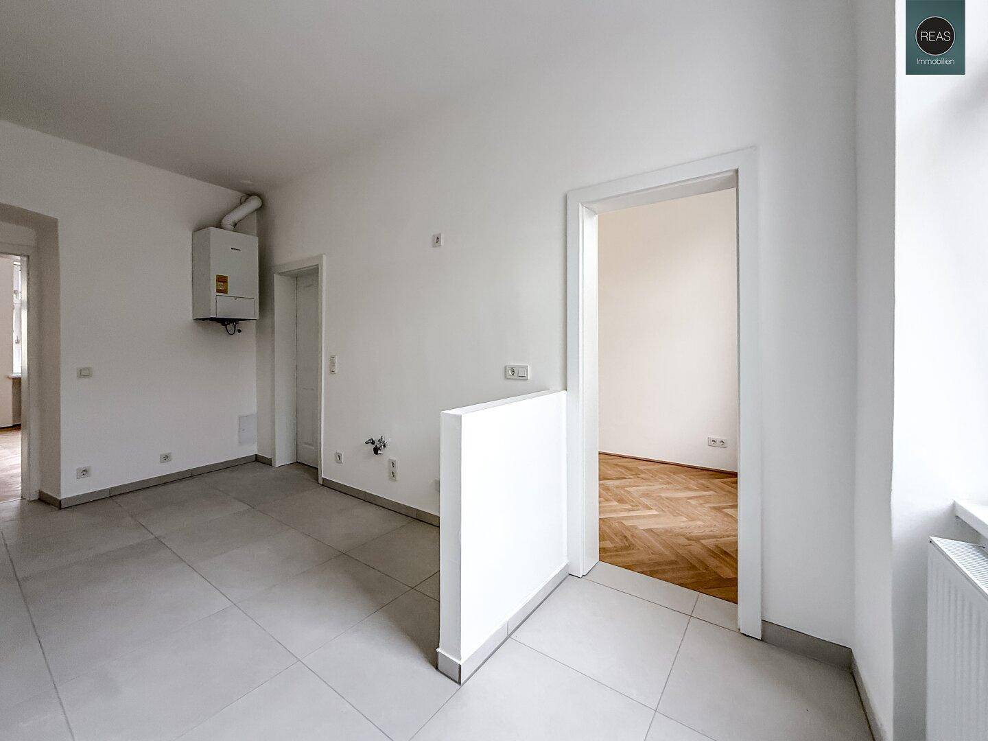 Raum mit grauen Fliesen und einem Wandheizkessel, vorbereitet für eine Kücheninstallation mit Zugang zu einem weiteren Zimmer.