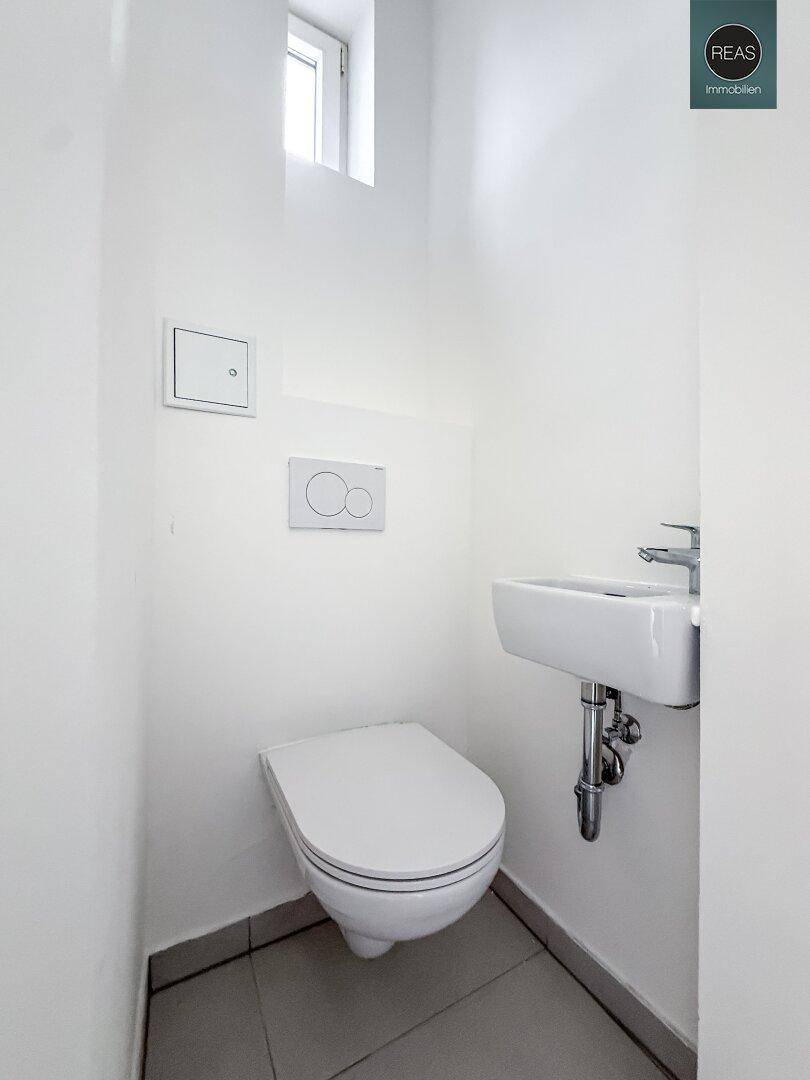 Kompaktes Gäste-WC mit wandhängender Toilette, kleinem Waschbecken und einem Fenster für natürliche Belüftung.