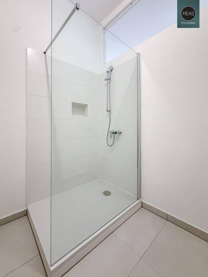 Geräumige, moderne Duschkabine mit Glasabtrennung und weißen Fliesen, integriert in ein Badezimmer mit grauen Bodenfliesen.