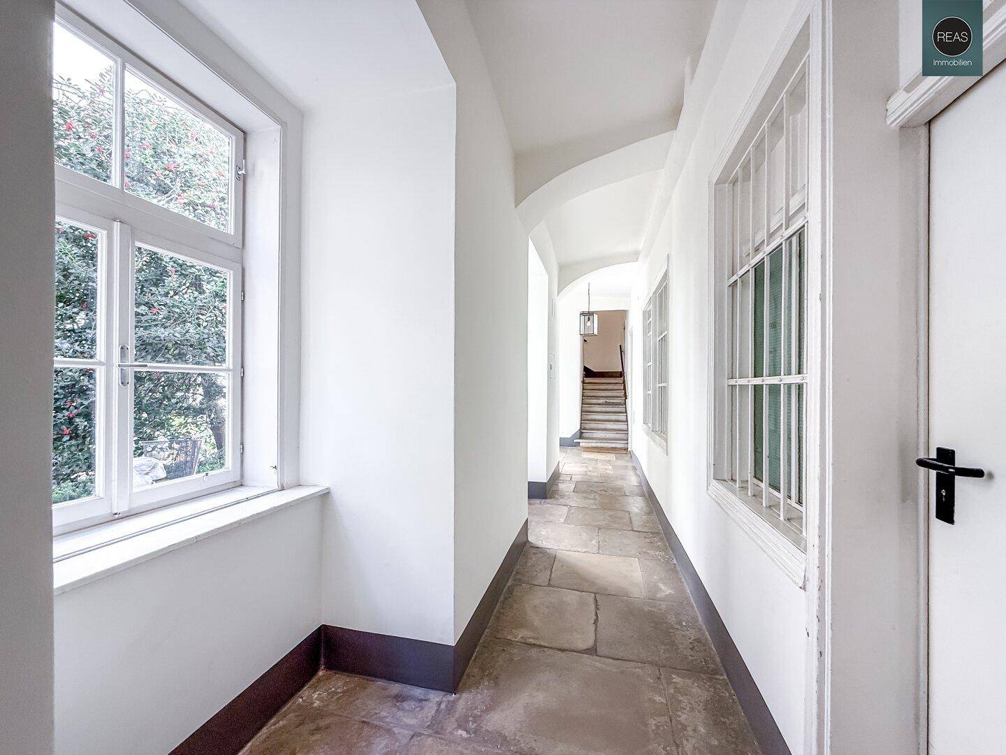 Langer Flur mit Steinboden, großen Fenstern und Blick auf eine Treppe im Hintergrund.