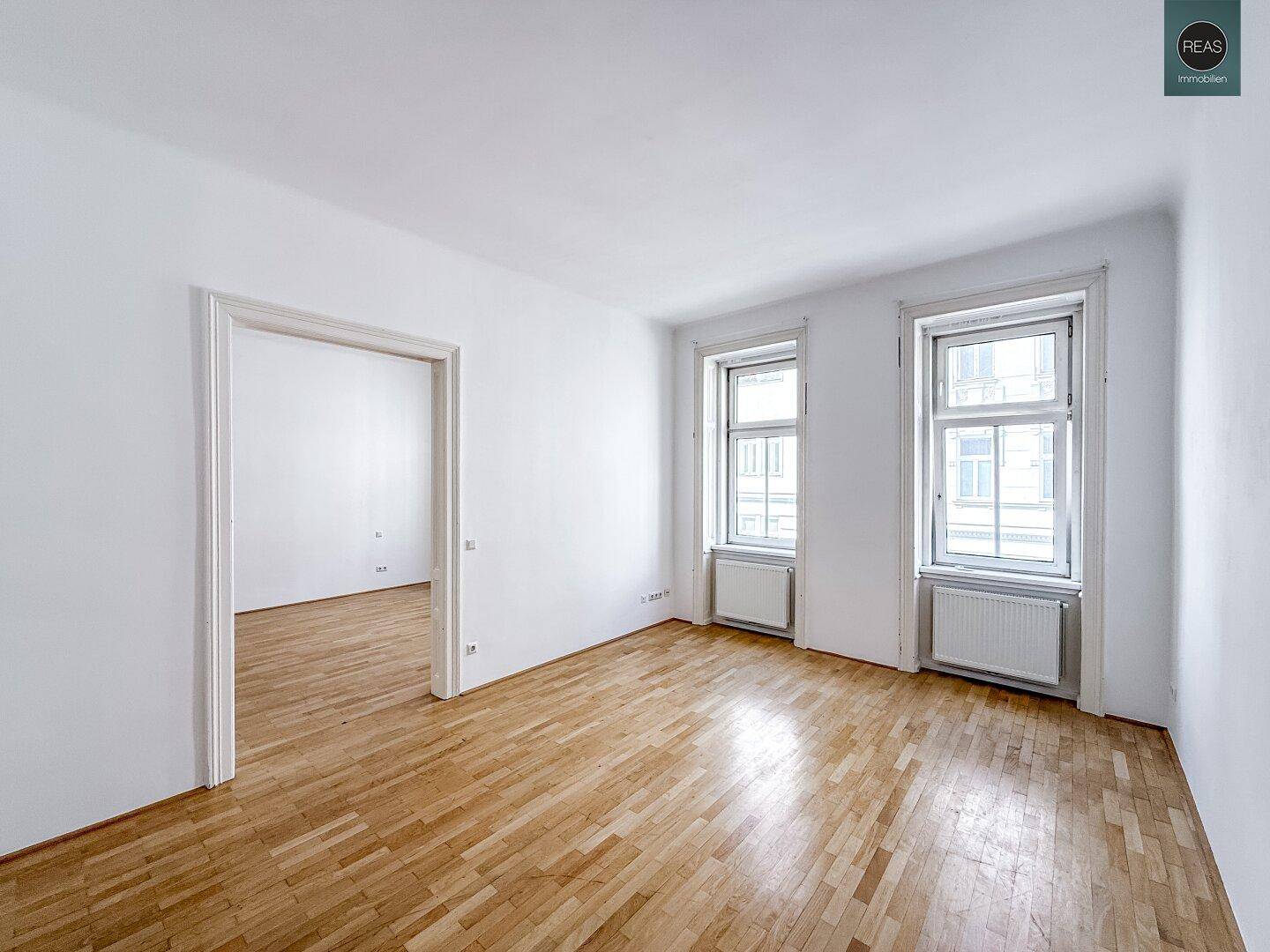 Geräumiger Wohnbereich mit Parkettboden und zwei hohen Fenstern, die viel Licht hereinlassen.