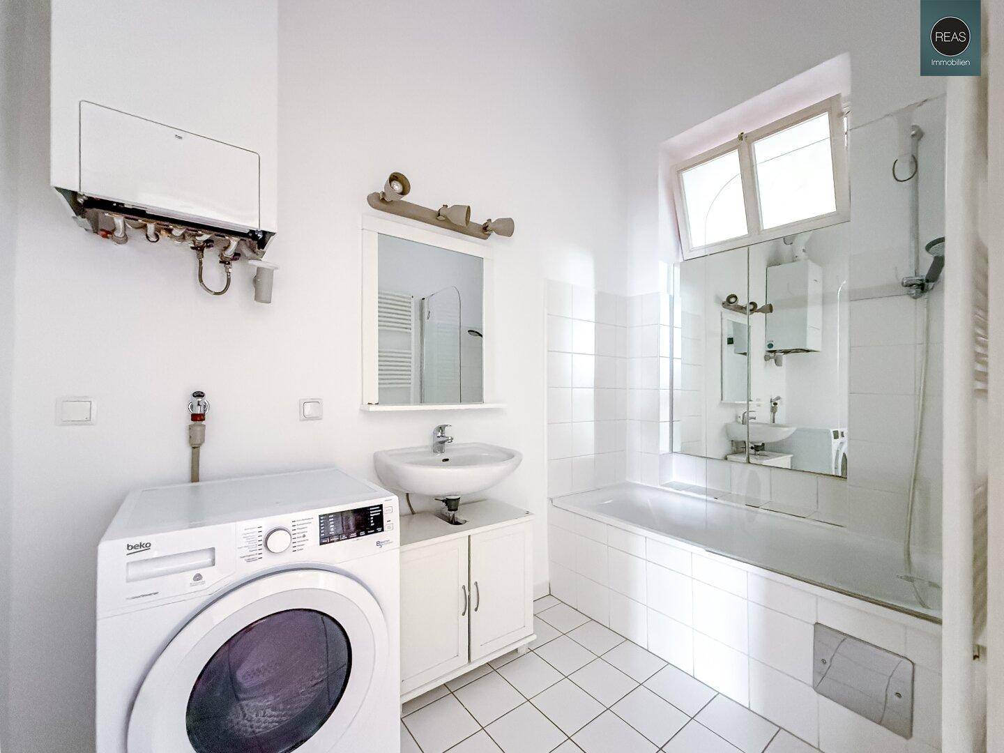 Helles Badezimmer mit Badewanne, Waschbecken, Waschmaschine und Fenster, weiß gefliest.