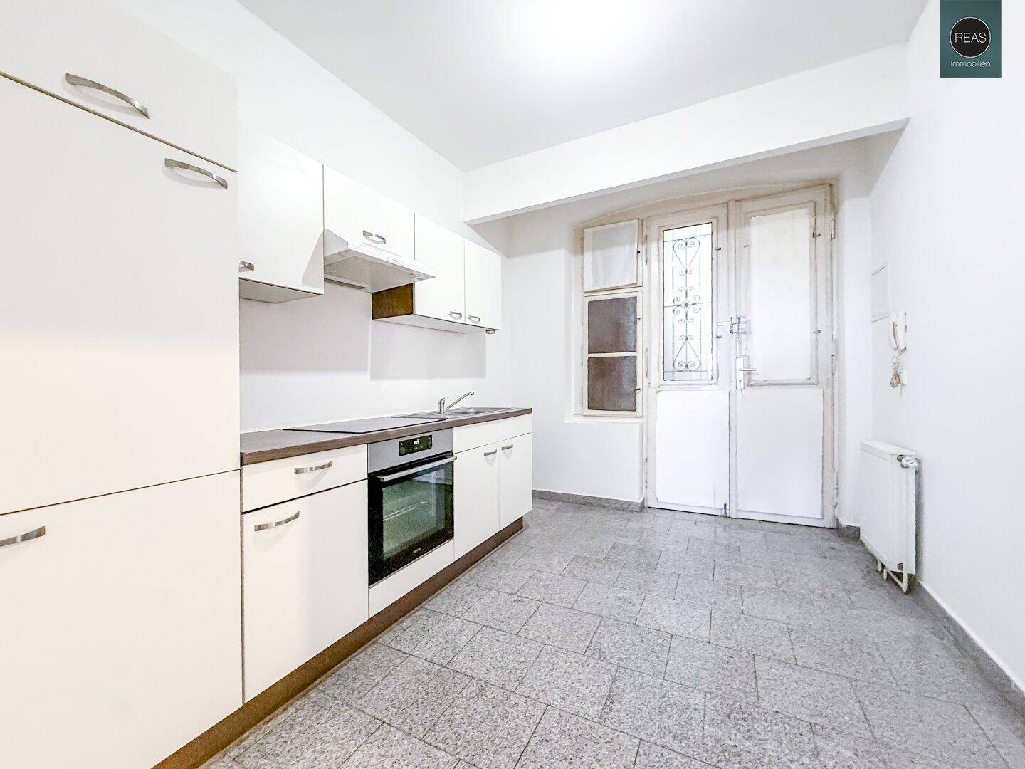 Einfache Küche mit weißen Schränken, integriertem Backofen und Fliesenboden, mit Fenster zur Außenseite.