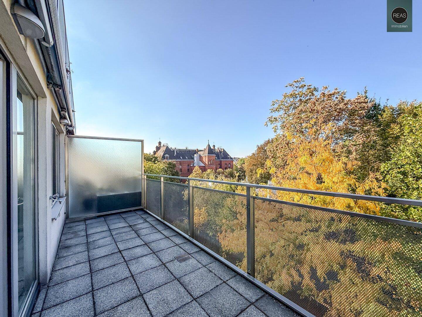 Großer Balkon mit Fliesenboden und weitem Blick auf grüne Bäume und ein historisches Gebäude.