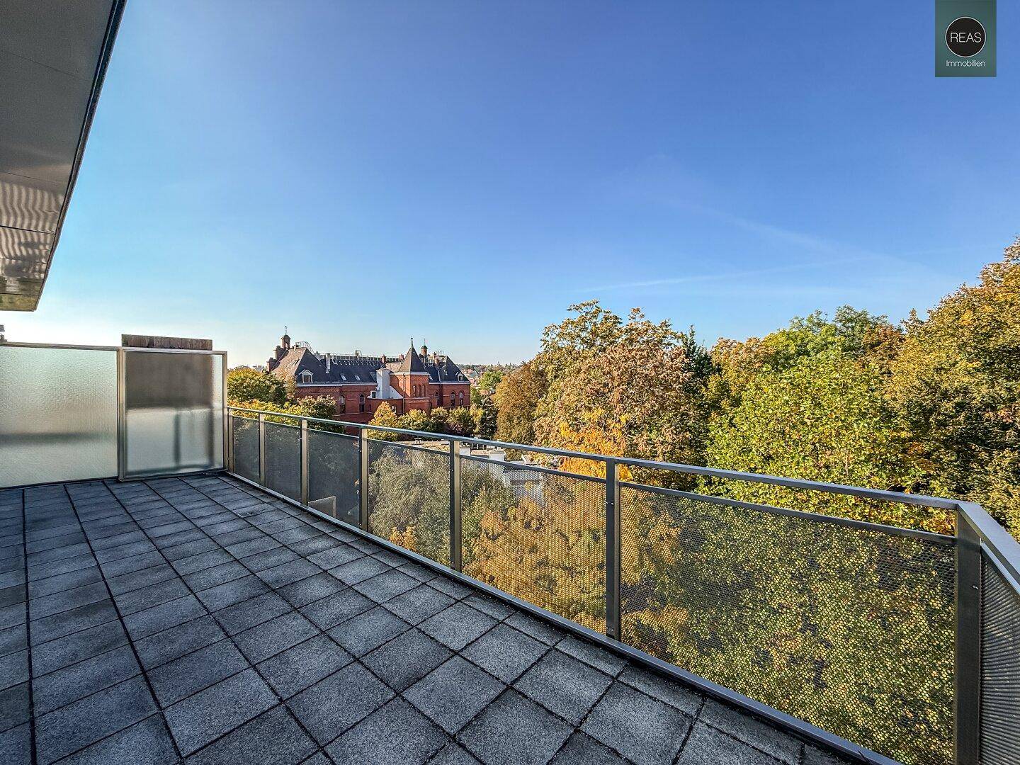 Großer Balkon mit Fliesenboden und weitem Blick auf grüne Bäume und ein historisches Gebäude.