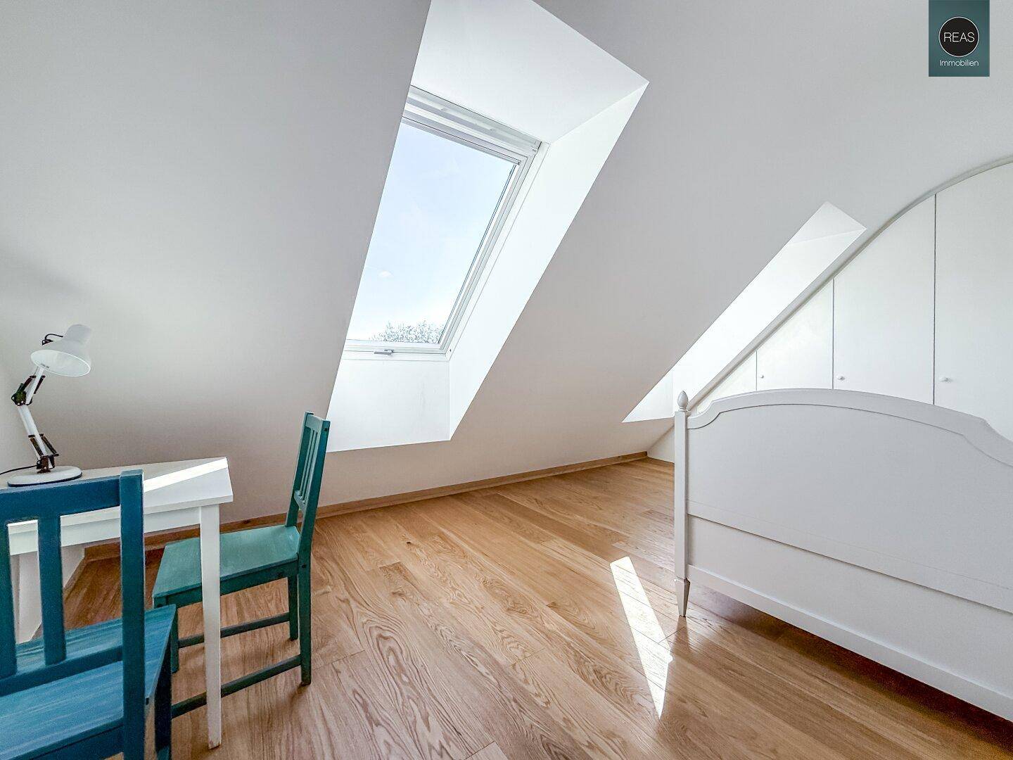 Helles Schlafzimmer im Dachgeschoss mit großem Dachfenster, Holzboden und Schreibtisch.