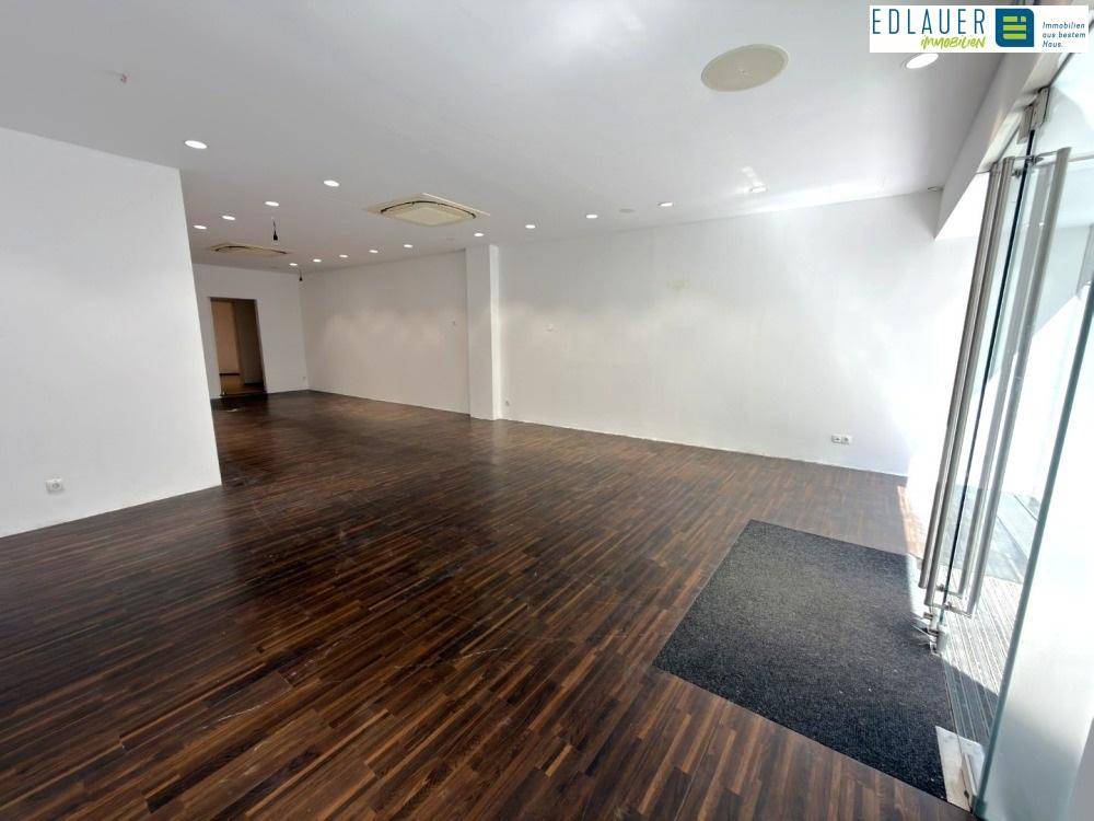 Heller Geschäftsraum mit dunklem Holzfußboden, weißen Wänden und großer Glasfront für viel Tageslicht.