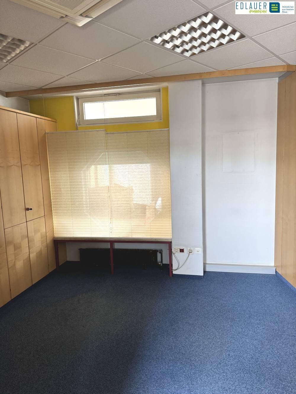 Kleinerer Büroraum mit Teppichboden, Einbauschränken und einem Fenster mit Jalousien.
