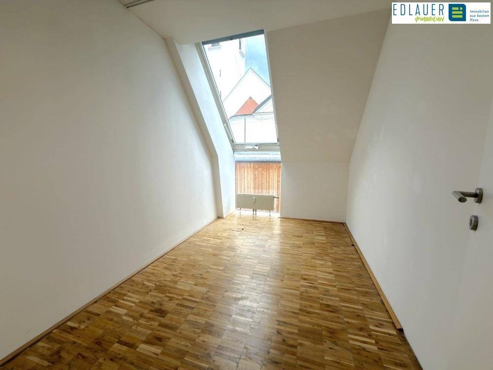 Heller Büroraum mit Holzparkett, weißen Wänden und einem schrägen Fenster, das viel Tageslicht hereinlässt.