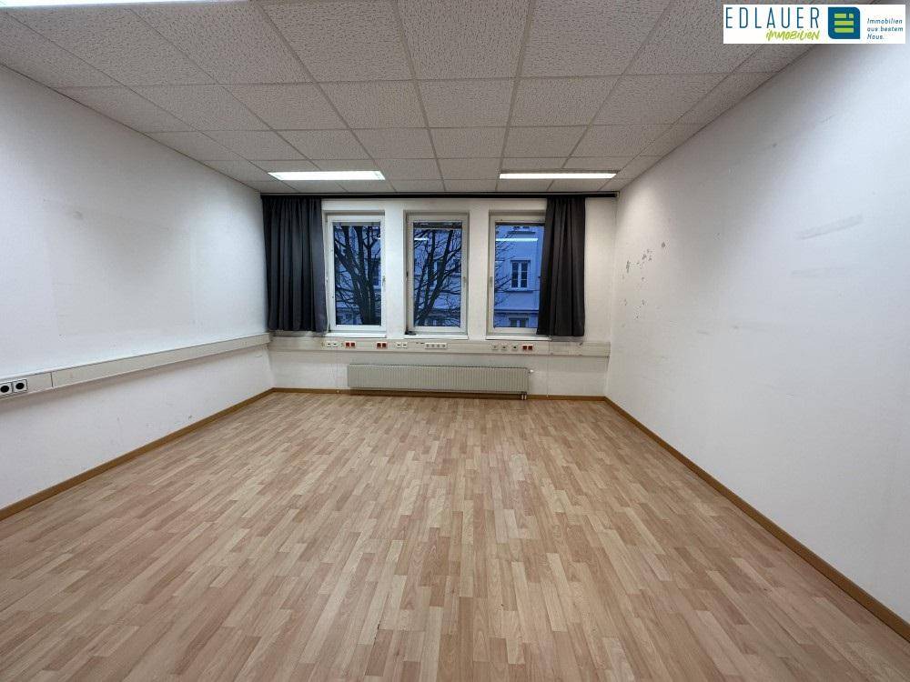 Geräumiger Büroraum mit hellem Laminatboden, drei Fenstern und abgehängter Decke, bereit zur Einrichtung.