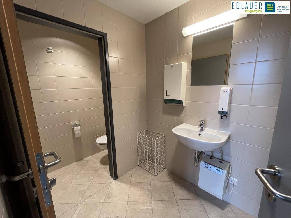 Saubere Toilette mit Fliesenboden, Waschbecken und Spiegel, ausgestattet mit Haltegriffen für Barrierefreiheit.