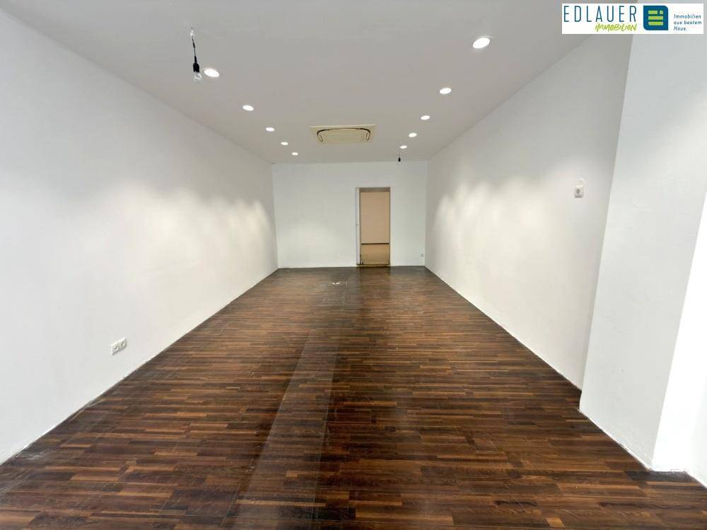 Langer, leerer Geschäftsraum mit dunklem Holzfußboden, weißen Wänden und Deckenbeleuchtung.