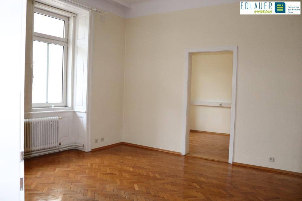 Heller Raum mit Fischgrätparkett und einem Durchgang zu einem weiteren Zimmer, typisch für einen Altbau.