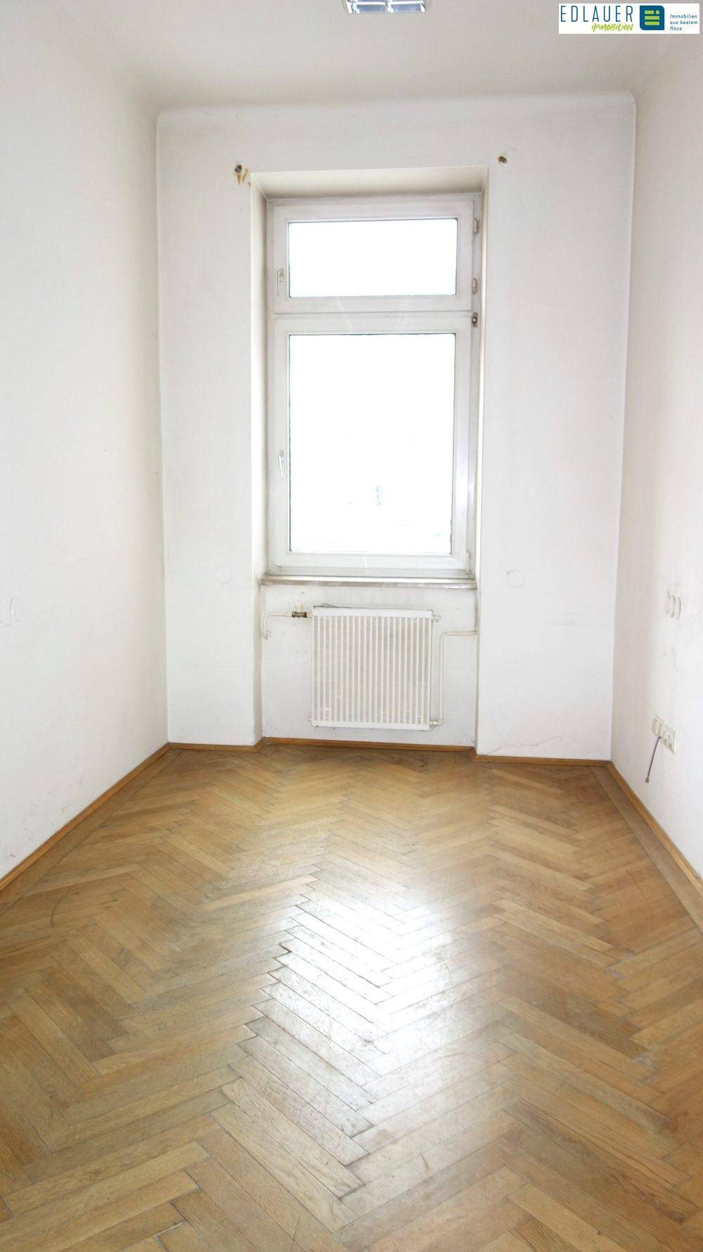 Heller, leerer Raum mit einem Fenster, Fischgrätparkett und einem Heizkörper unter der Fensterbank.