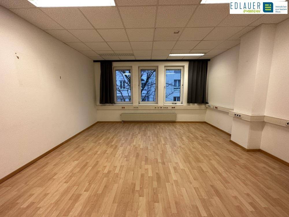 Heller Büroraum mit Laminatboden, drei Fenstern und weißen Wänden, bietet eine angenehme Arbeitsatmosphäre.