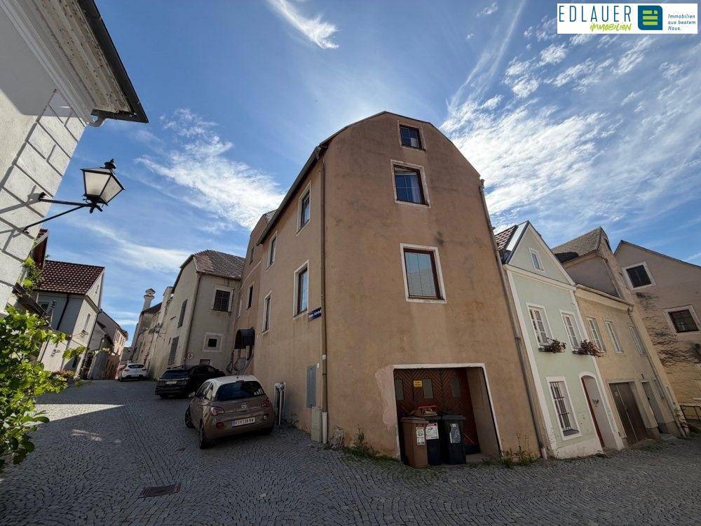 Gebäudeansicht an einer gepflasterten Straße mit angrenzenden Häusern und blauem Himmel.