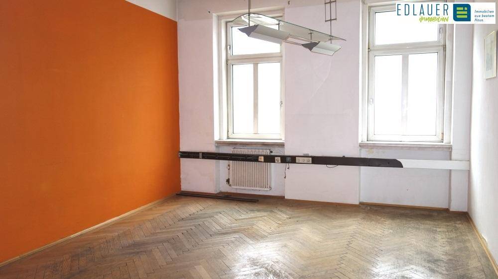 Geräumiger Raum mit orangefarbener Wand, Fischgrätparkett und zwei großen Fenstern.