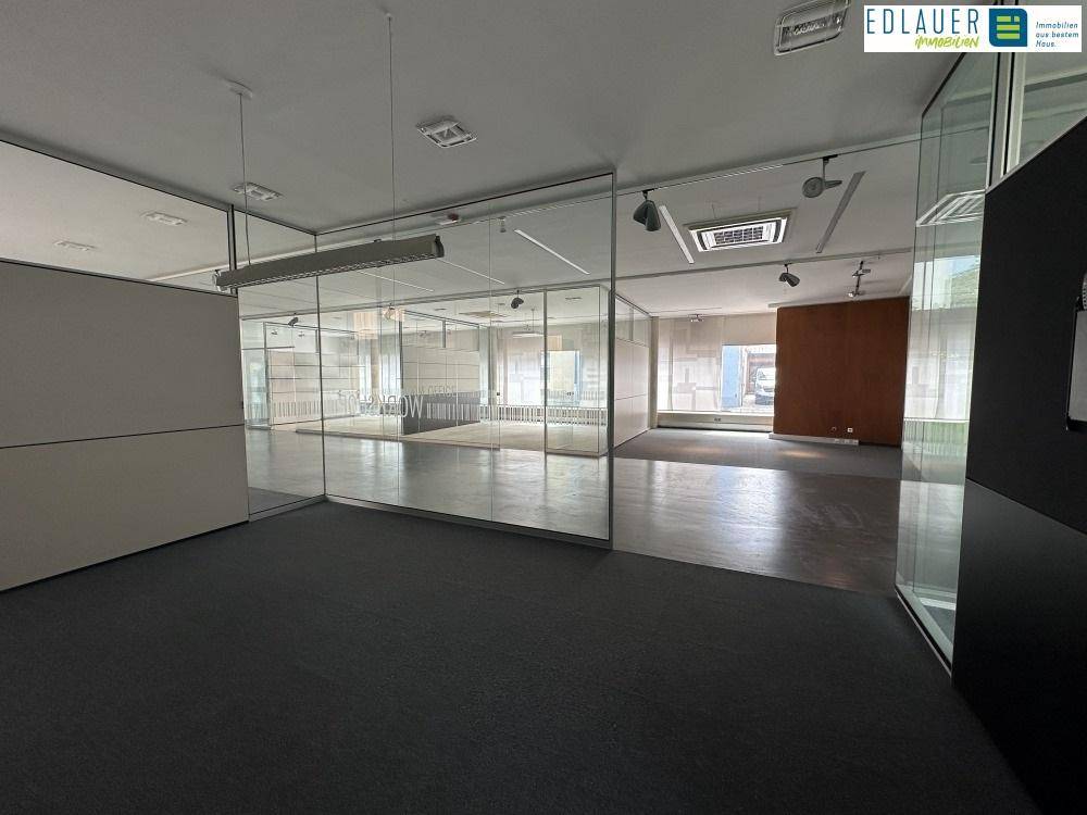 Offener Bürobereich mit Glaswänden, dunklem Teppichboden und Blick auf weitere, helle Büroräume.