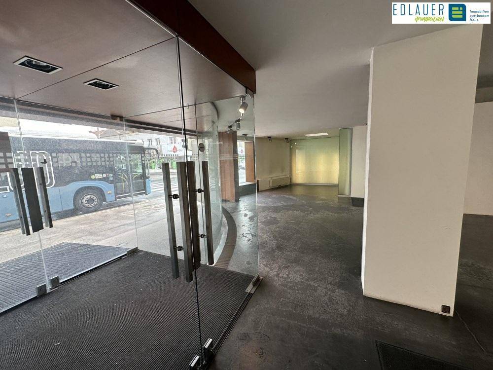 Eingangsbereich eines Geschäfts mit großen Glastüren, dunklem Boden und Blick auf die Straße mit einem Bus.