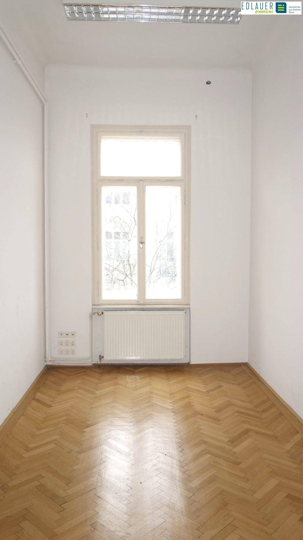 Kleiner, heller Raum mit Fischgrätparkett, einem Fenster und Deckenleuchten, ideal als Büro.