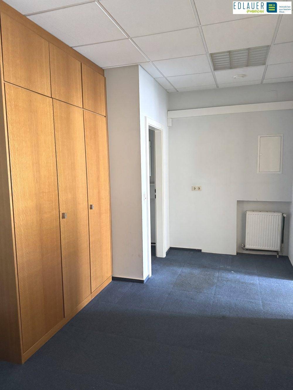 Büroraum mit blauem Teppichboden, Einbauschränken und einem Heizkörper unter dem Fenster.